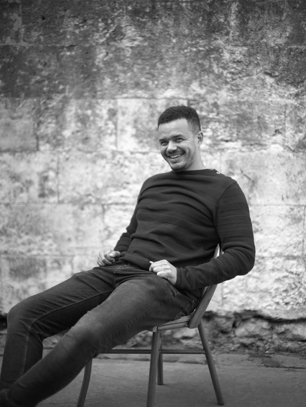 Danny Morris smiling, sat on a chair in front of a stone wall, black and white